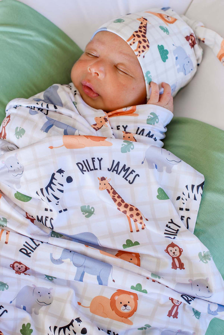 Close-up of a sleeping newborn with a hand by his face, swaddled in a Caden Lane personalized Wild Child swaddle reading "Riley James," paired with a matching knot hat.