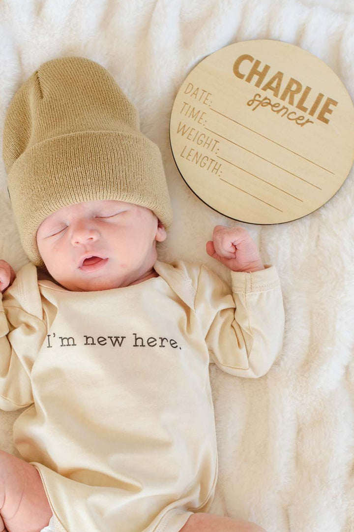 Sleeping newborn in Caden Lane's "I'm new here" cream bodysuit and tan knit beanie, lying beside a Caden Lane personalized wooden birth announcement sign reading "Charlie Spencer" with blank date, time, weight, and length lines.