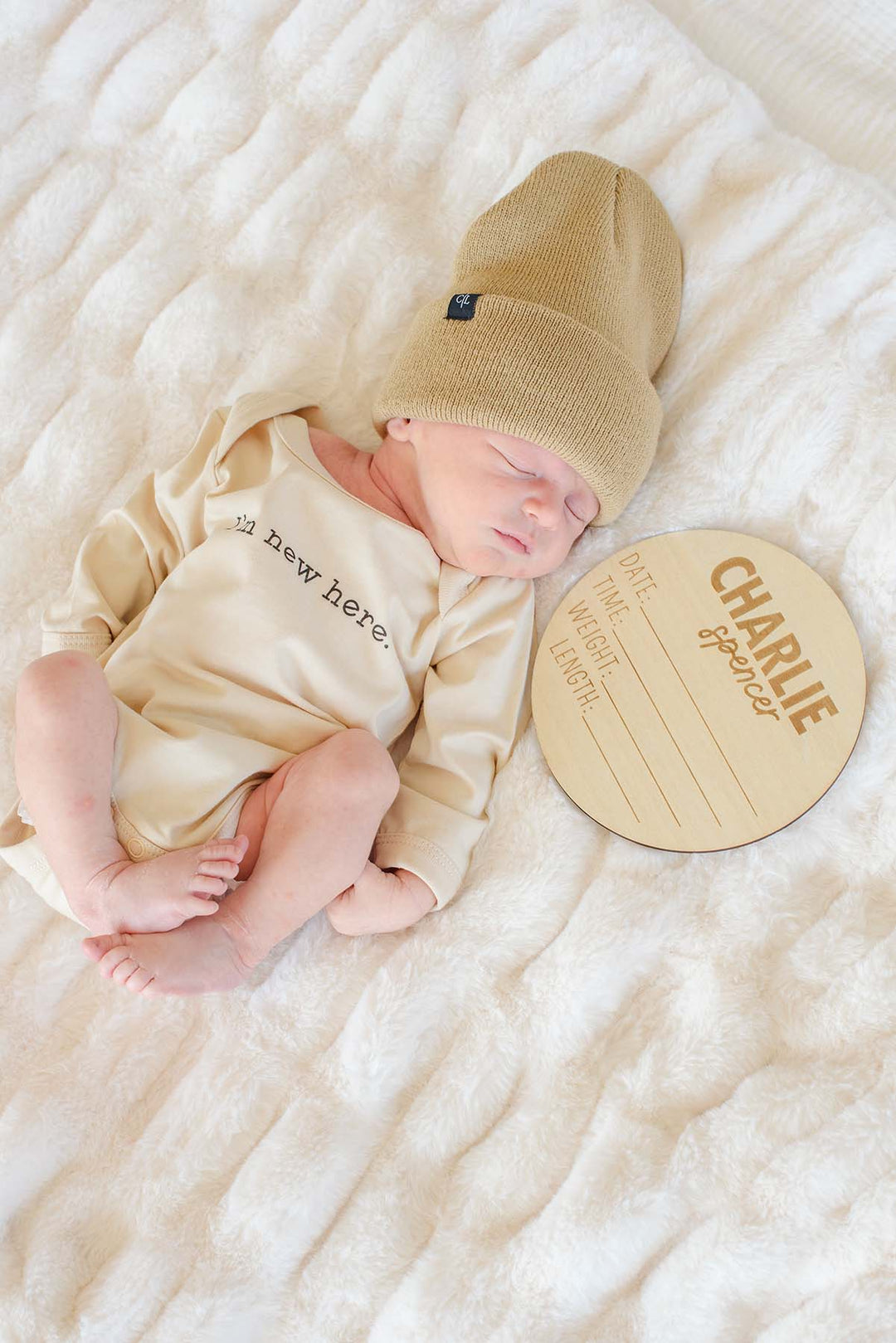 Sleeping newborn wearing a Caden Lane tan knit beanie with logo tag and cream "I'm new here" bodysuit, lying next to a personalized wooden "Charlie Spencer" name sign on a fuzzy white blanket.
