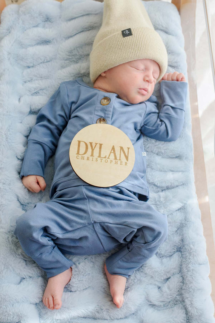 Close-up overhead view of a sleeping newborn in a Caden Lane tan knit beanie and blue button-shoulder outfit, with a Caden Lane wooden "Dylan Christopher" sign resting on his chest.