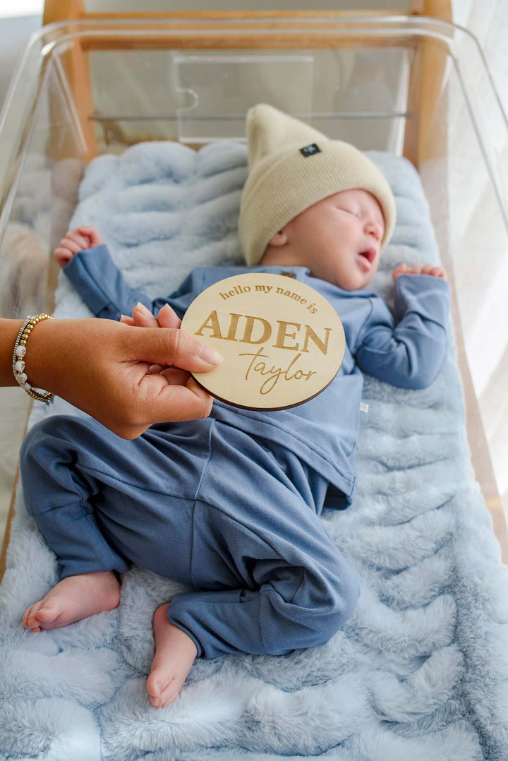 A hand with a beaded bracelet holds a Caden Lane wooden "hello my name is Aiden Taylor" sign above a sleeping newborn boy in a Caden Lane tan knit beanie and blue button-detail outfit.