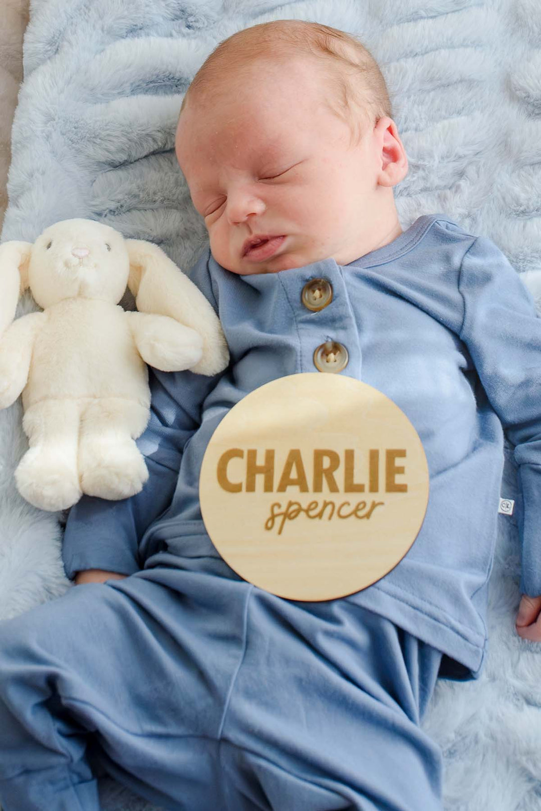 Sleeping newborn boy in a Caden Lane blue button-shoulder sleeper, lying beside a plush bunny with a Caden Lane engraved wood sign reading "Charlie Spencer" resting on his outfit.