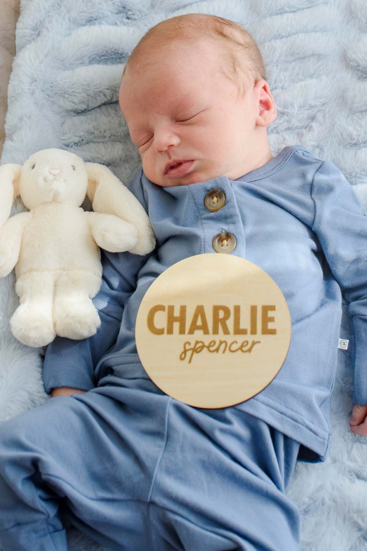 Sleeping newborn boy in a Caden Lane blue button-shoulder sleeper, lying beside a plush bunny with a Caden Lane engraved wood sign reading "Charlie Spencer" resting on his outfit.