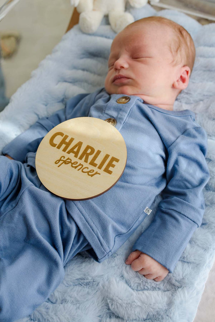 Close-up of a sleeping newborn in a Caden Lane blue button-shoulder sleeper with a Caden Lane logo tag, holding a wooden "Charlie Spencer" birth announcement sign against his chest.