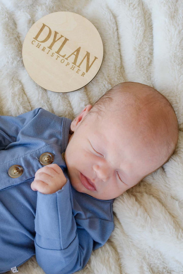 Close-up of a sleeping newborn in a Caden Lane blue button-detail outfit, resting on cream faux fur next to a Caden Lane engraved wood sign reading "Dylan Christopher."