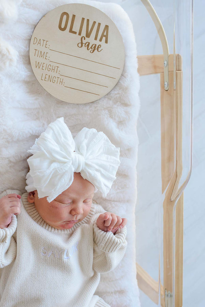 Close-up of a sleeping newborn in a Caden Lane white bow headband and cream sweater, positioned below a wooden "Olivia Sage" birth announcement plaque in a clear hospital bassinet.