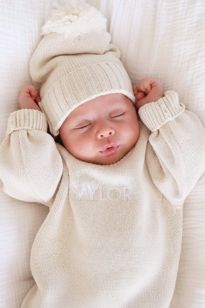 long sleeve beige personalized sweater romper for babies embroidered with name with matching pom pom beanie 