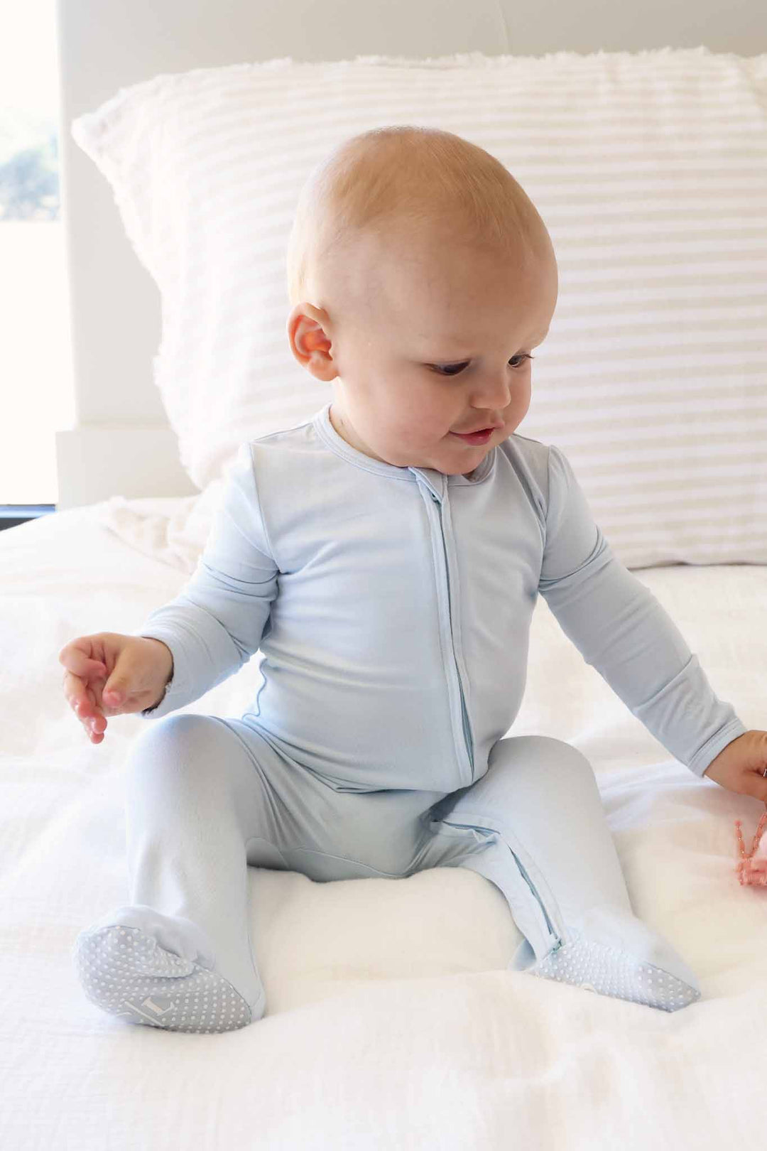 baby boy sitting on bed wearing a light blue zipper footie pajama with foot grips 