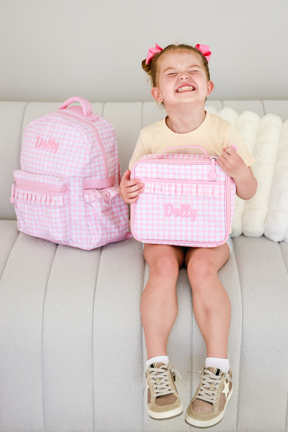 girl sitting on the couch holding a pink gingham embroidered lunchbox with the matching backpack 