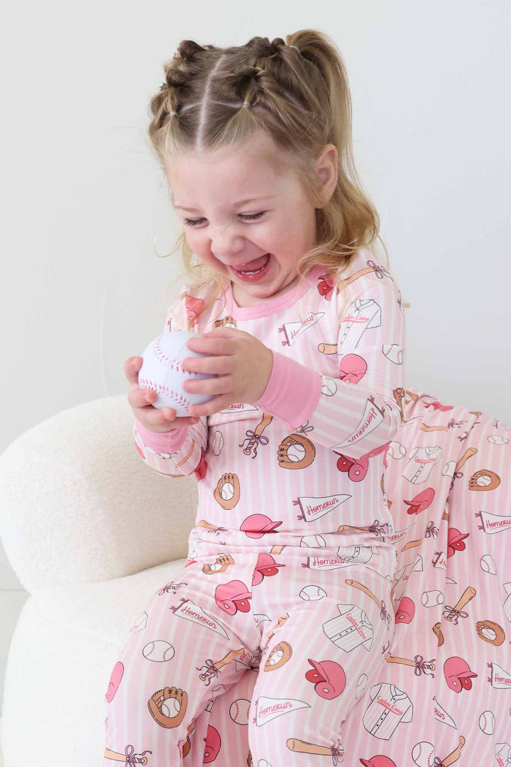 girl sitting on chair wearing a pink and white striped two piece baseball themed pajama set holding a foam baseball 