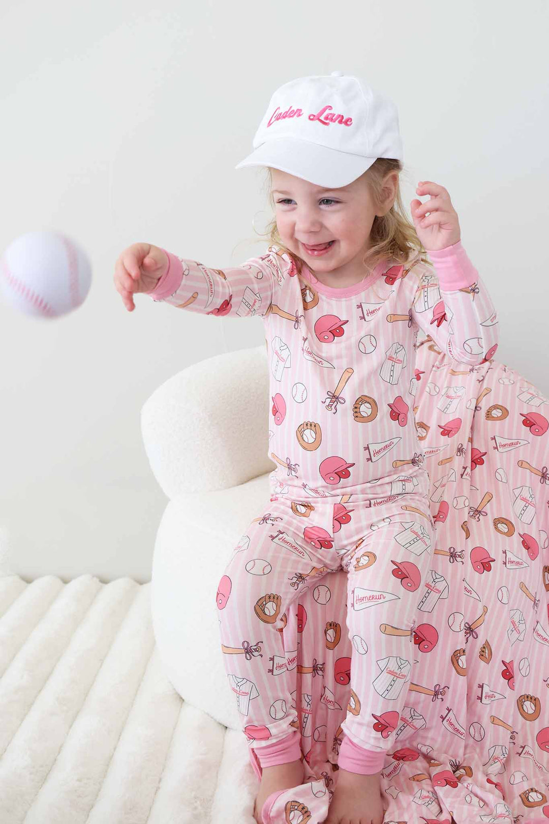 girl sitting on chair wearing the ballpark babes two piece pajama set with an embroidered caden lane baseball cap throwing a baseball 