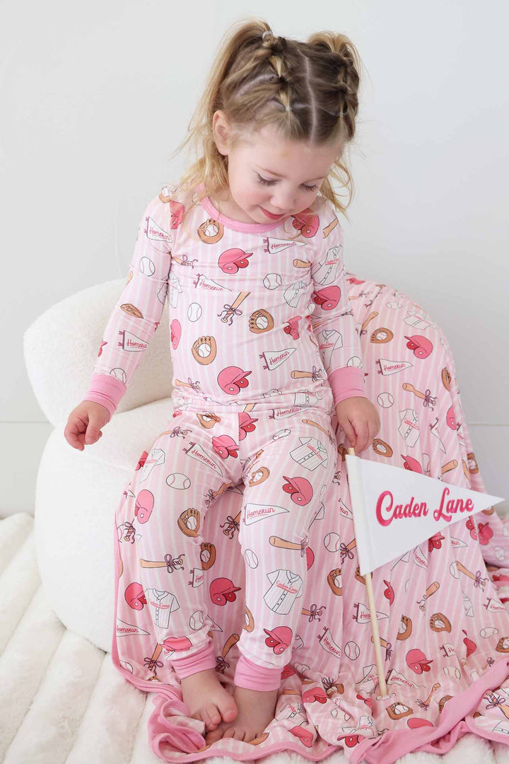 girl sitting on white chair in pink girly baseball themed two piece pajama set next to white caden lane flag banner and matching bamboo blanket 
