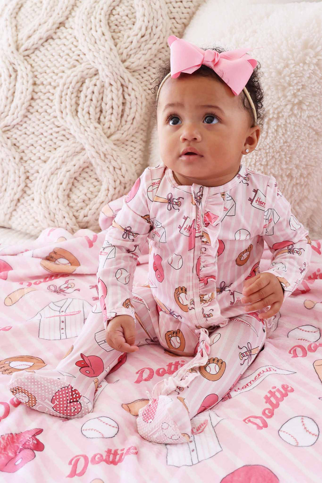 baby girl sitting on bed in ballpark babes bamboo ruffle zipper footie with matching pink ribbon bow headband 