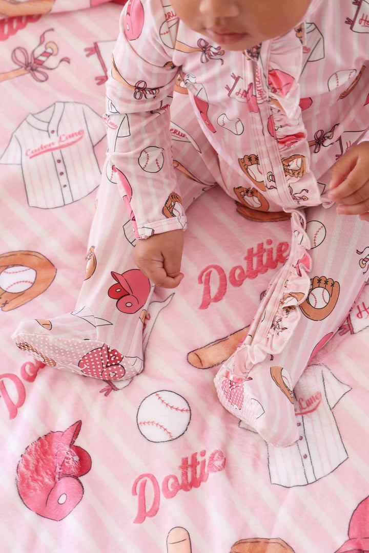top view of baby girl sitting on pink baseball themed personalized blanket wearing a matching ruffle zipper footie 