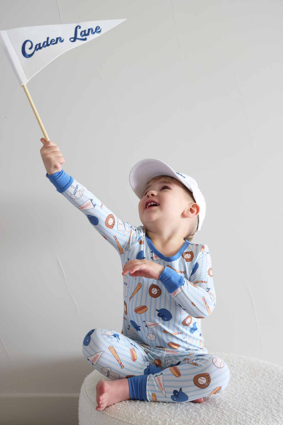 boy wearing ballpark bros bamboo two piece pajama set with matching baseball cap and holding up a flag
