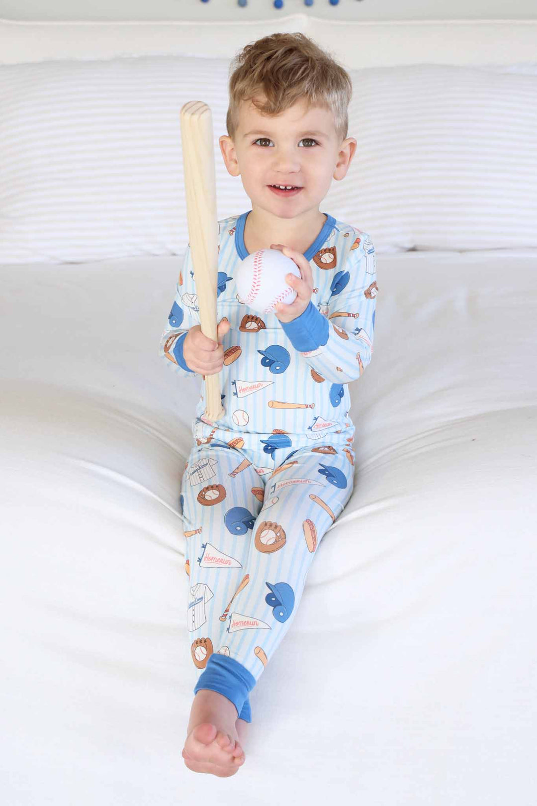 boy sitting on bed in ballpark bros two piece pajama set holding a baseball bat and ball