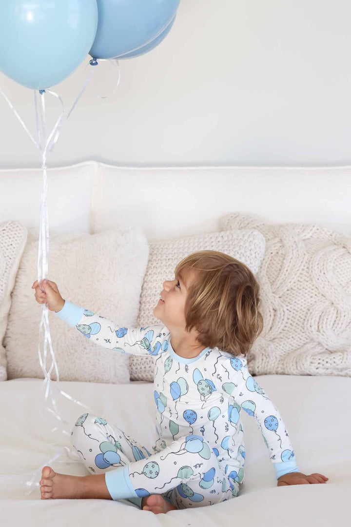 boy sitting on bed holding and looking up at balloons in blue and green balloon pajama set 