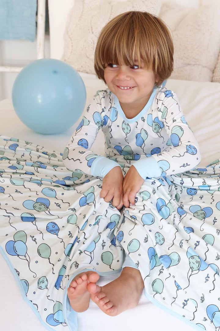 boy sitting on bed wearing blue and green balloon pajamas with a matching blanket draped over him 