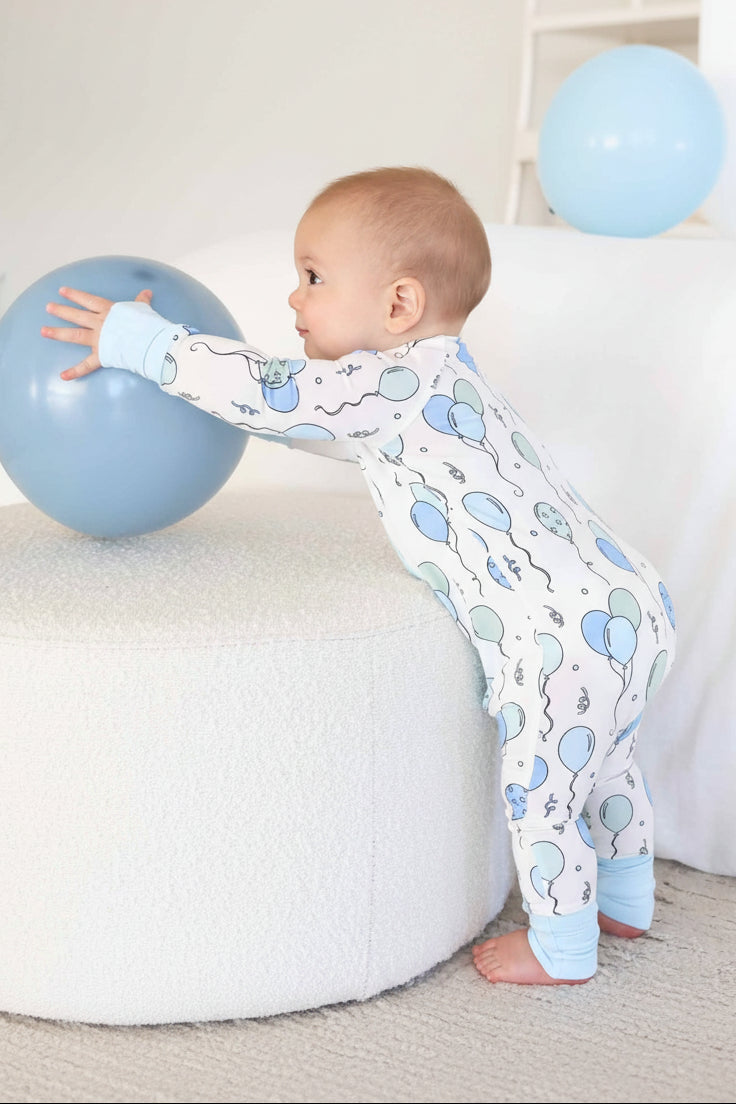 standing boy wearing balloon and streamer pajama romper reaching for a blue balloon
