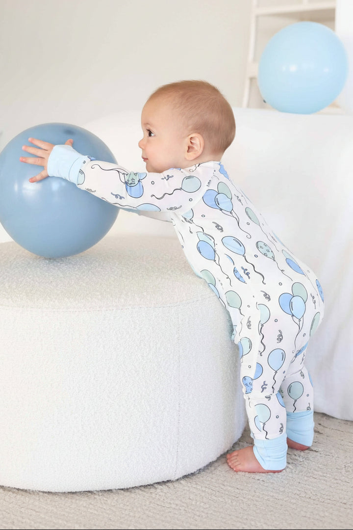 standing boy wearing balloon and streamer pajama romper reaching for a blue balloon