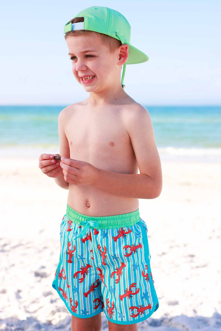 boy standing on the beach in blue and green striped lobster swim trunks 