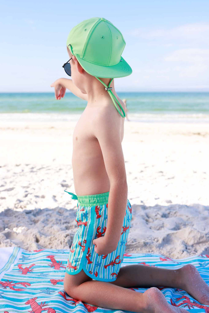 boy kneeling on towel at the beach in lobster swim trunks with a matching green surf hat 