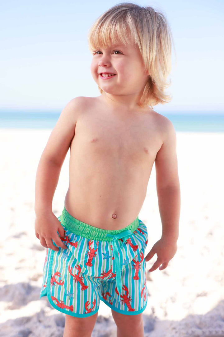 boy standing on the beach in striped lobster swim trunks 