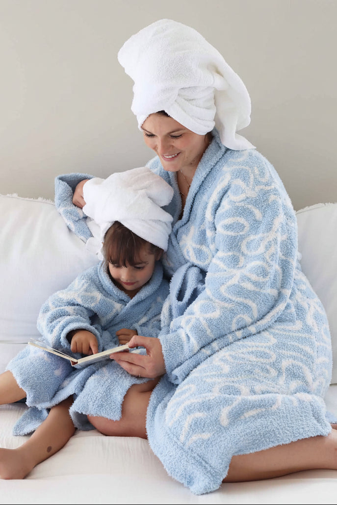 mom and daughter sitting on a couch reading a book wearing matching plush robes that are blue with white bows with towels wrapped on their heads 