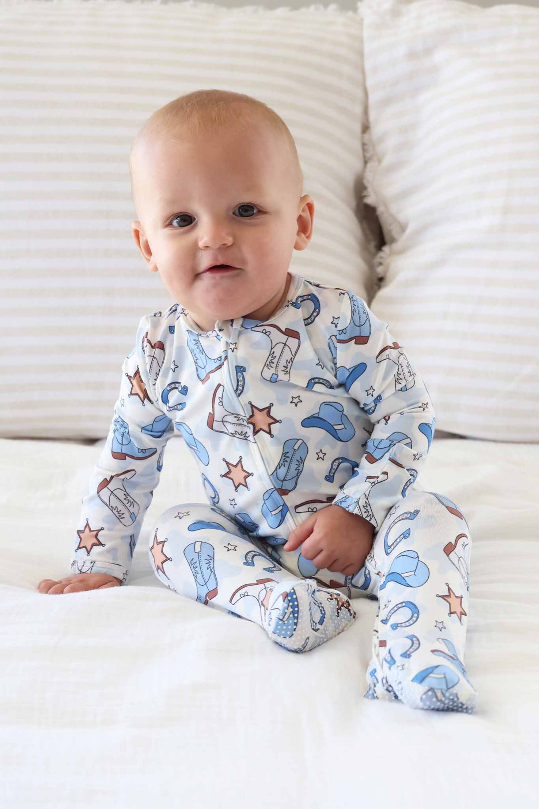 sitting baby boy wearing a footie pajama with blue cowboy hats, boots, horseshoes and sherriff stars