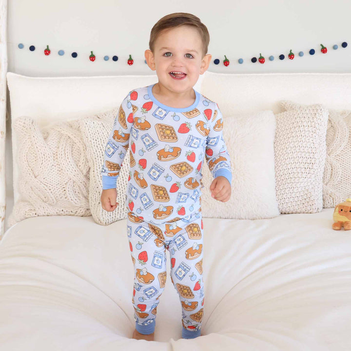 boy standing on a bed wearing a two piece pajama set with pancakes, waffles, fruit and jelly on them