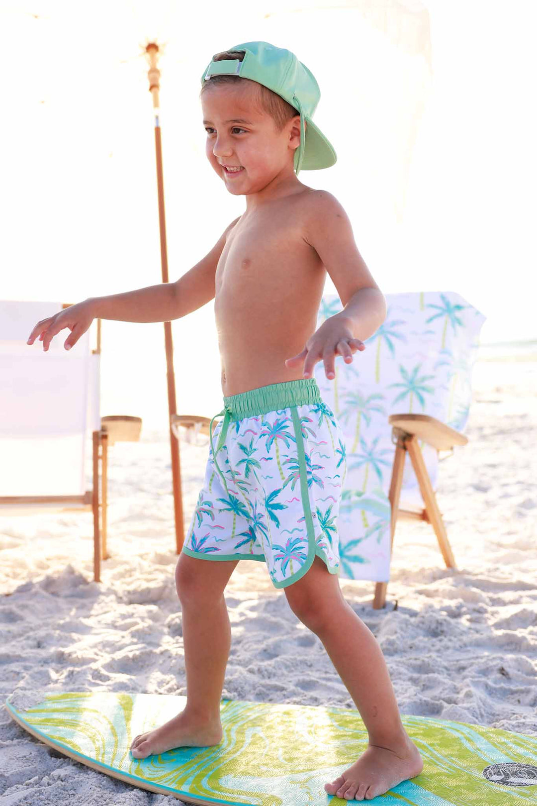boy standing on surf board in neon palm tree swim trunks with a backwards green hat