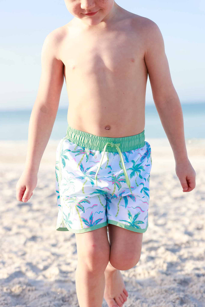 boy walking on beach in blue, green and pink palm tree swim trunks 