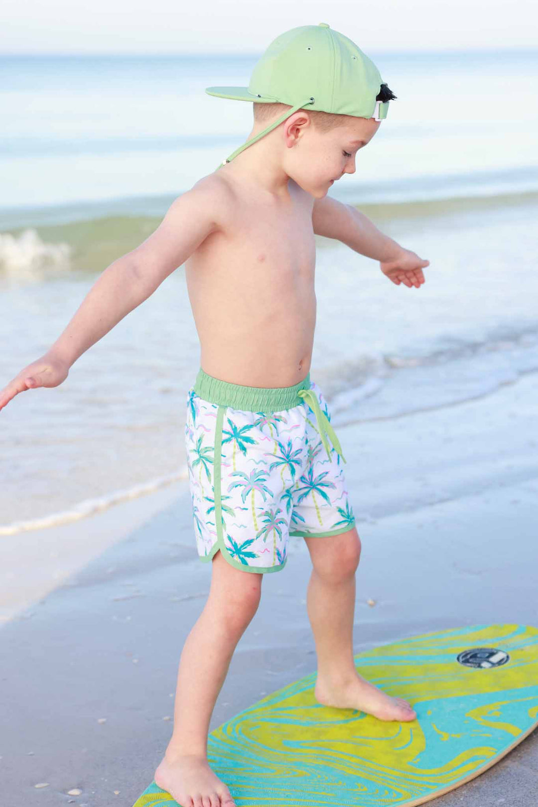 boy standing on surf board in neon palm tree swim trunks 