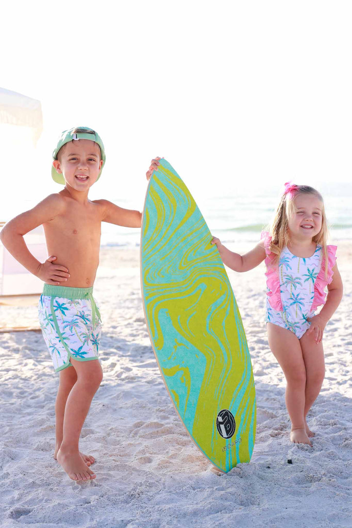 brother and sister standing on beach wearing neon palm tree swimsuits holding up a green and blue surf board 