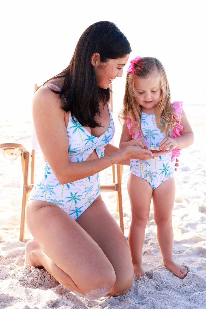 mom wearing cutout one piece palm tree swimsuit and looking at star fish on beach with daughter in matching ruffle one piece 