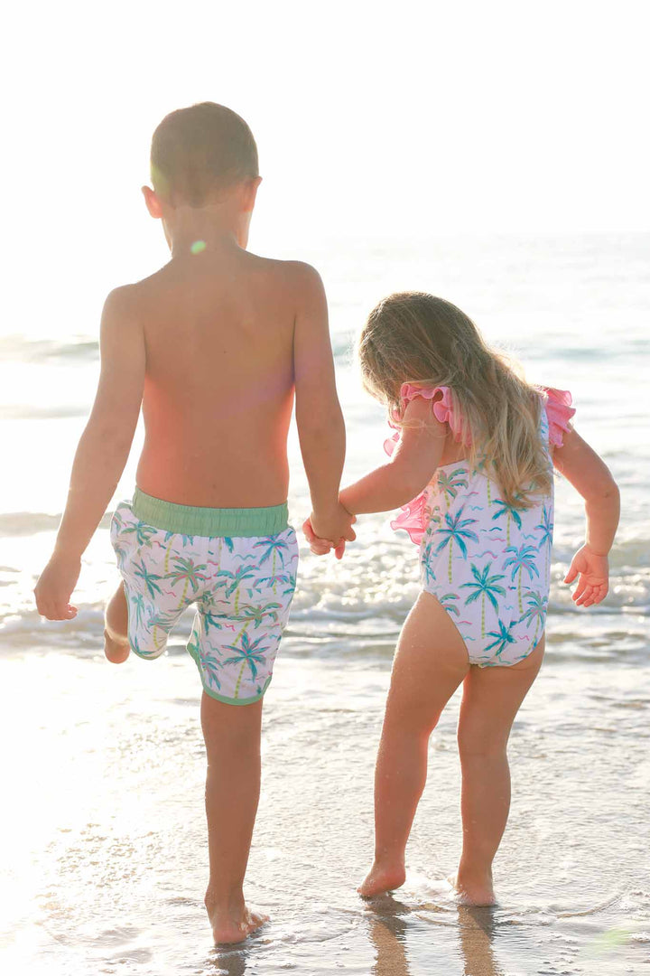 brother and sister wearing matching palm tree swimsuits and holding hands on the beach