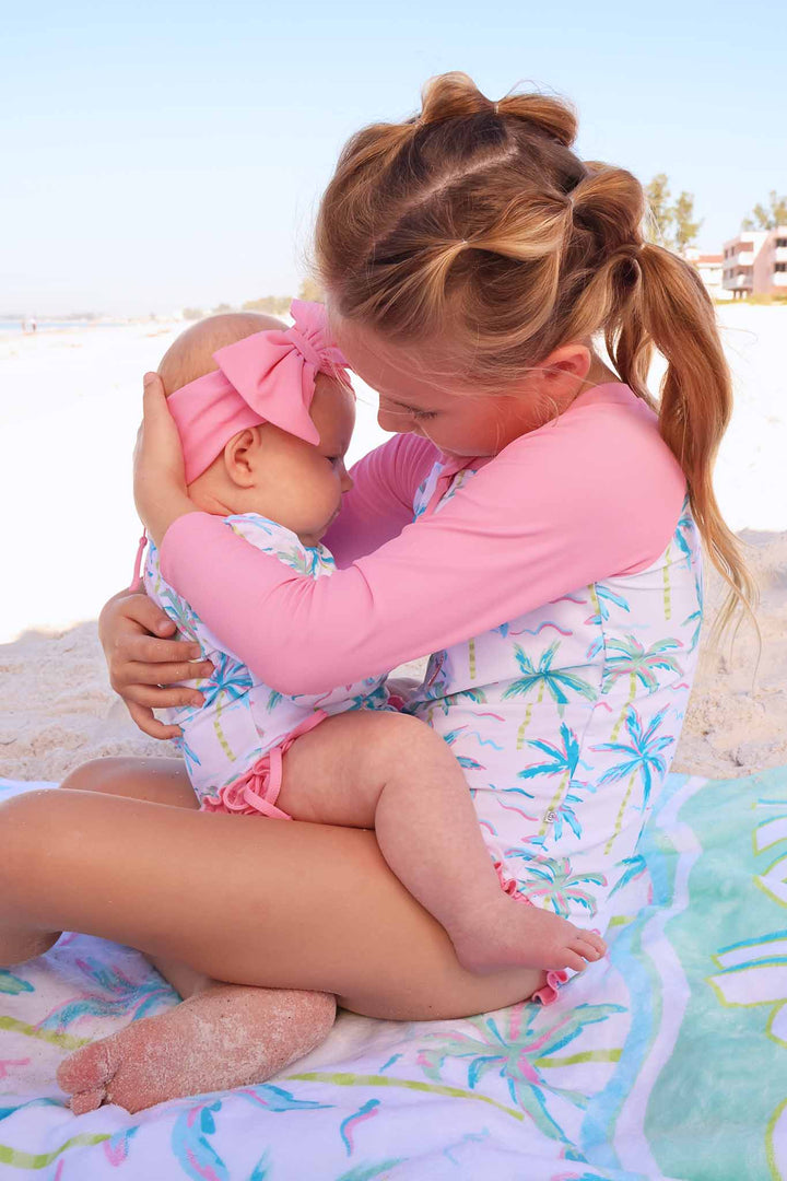 girl holding baby sitting on beach towel in matching palm tree swimsuits 