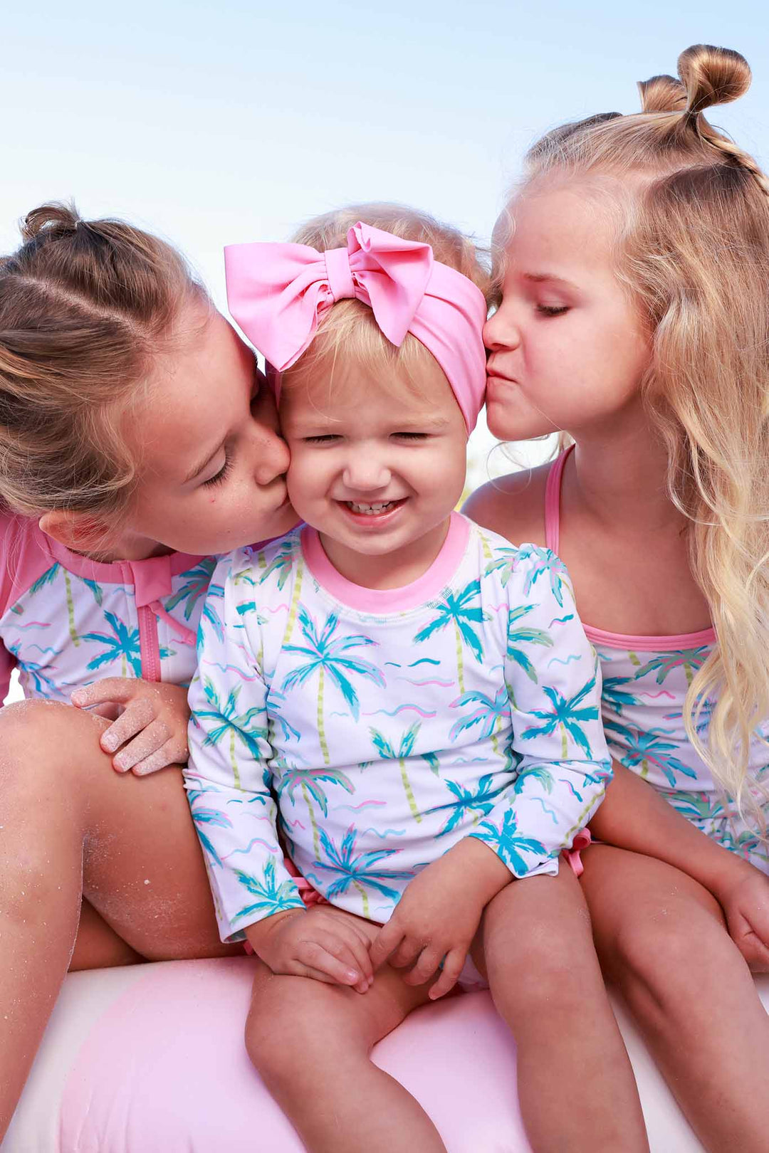 sisters sitting on innertube wearing matching palm tree swimsuits and giving the baby kisses on the cheek
