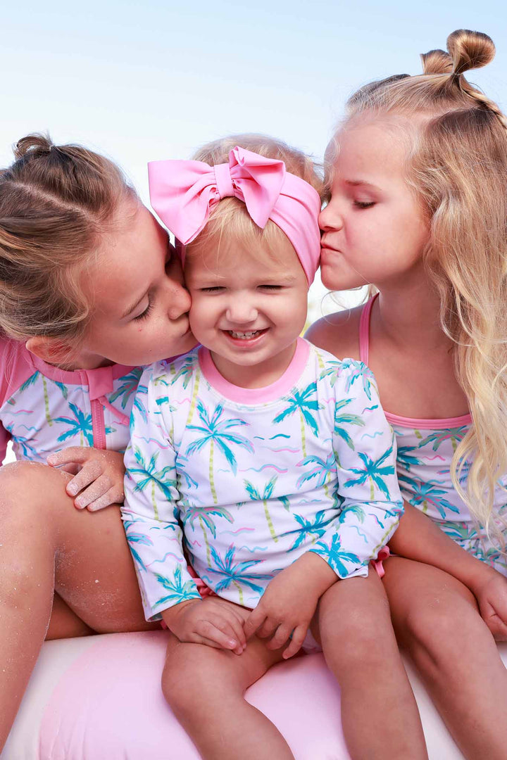 sisters sitting on innertube wearing matching palm tree swimsuits and giving the baby kisses on the cheek