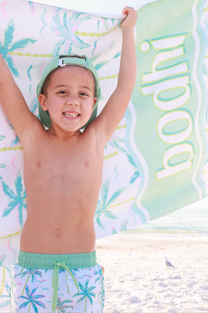 boy standing on beach in palm tree swim trunks and backwards green surf hat with personalized towel behind him 