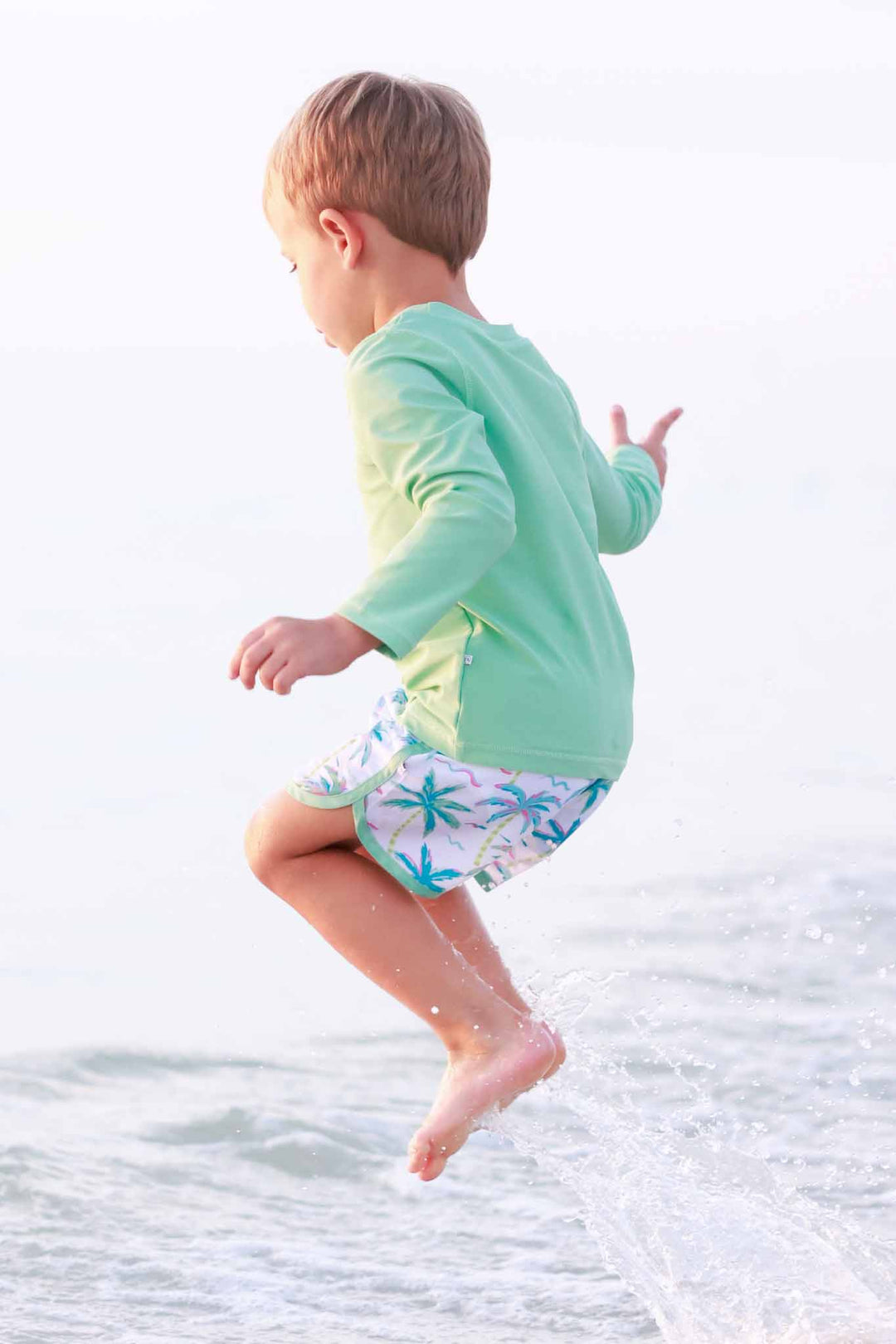 boy jumping in ocean wearing a neon green long sleeve rash guard shirt with colorful palm tree swim trunks 