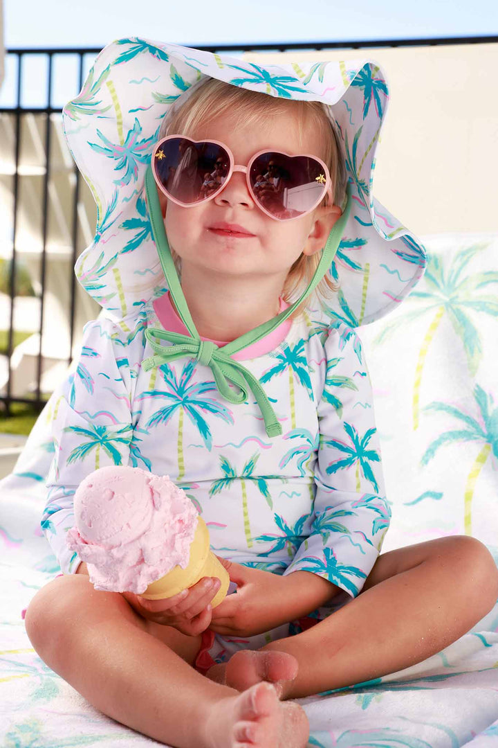 girl sitting on beach chair in palm tree long sleeve rash guard with ruffle bottom wearing a matching hat and heart sunglasses holding ice cream 