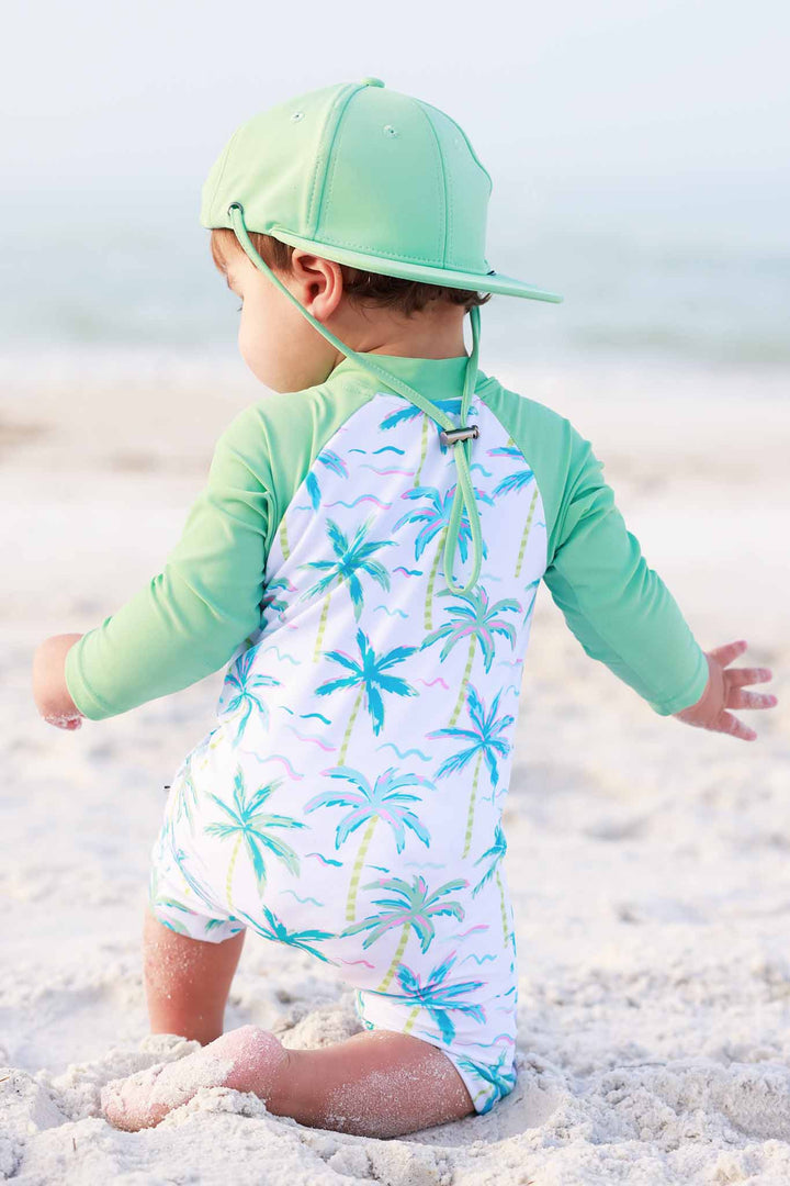baby boy kneeling on sand wearing long sleeve rash guard shortie romper with neon palm trees
