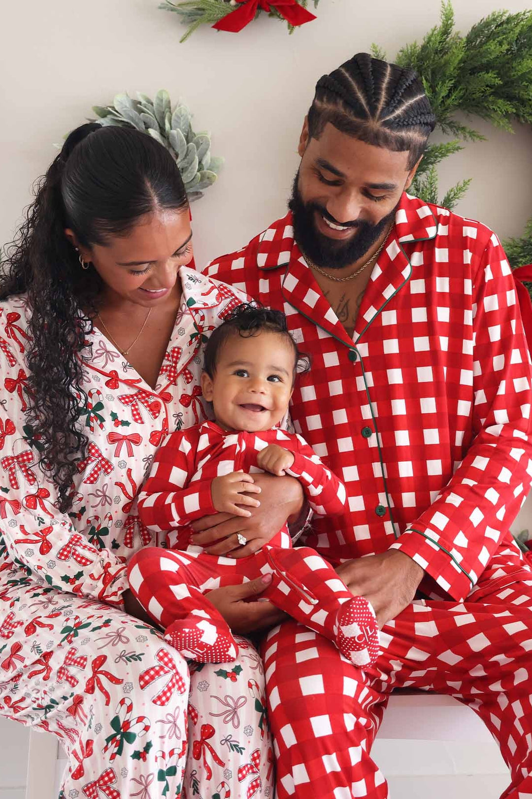 Smiling family wearing coordinating Caden Lane Christmas pajamas, with parents holding their baby in matching red and white checkered holiday sleepwear
