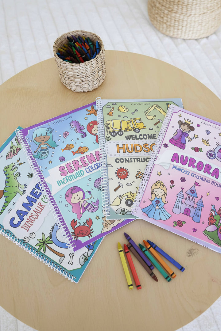 Four personalized Caden Lane coloring books – Cameron's Dinosaur, Serena's Mermaid, Hudson's Construction, and Aurora's Princess – fanned out on a wood table with a basket of crayons and loose crayons