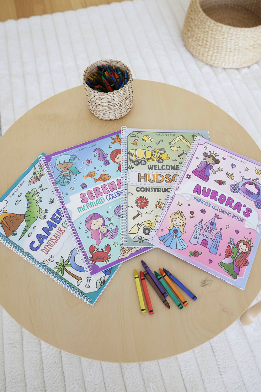 Four personalized Caden Lane coloring books – Cameron's Dinosaur, Serena's Mermaid, Hudson's Construction, and Aurora's Princess – fanned out on a wood table with a basket of crayons and loose crayons