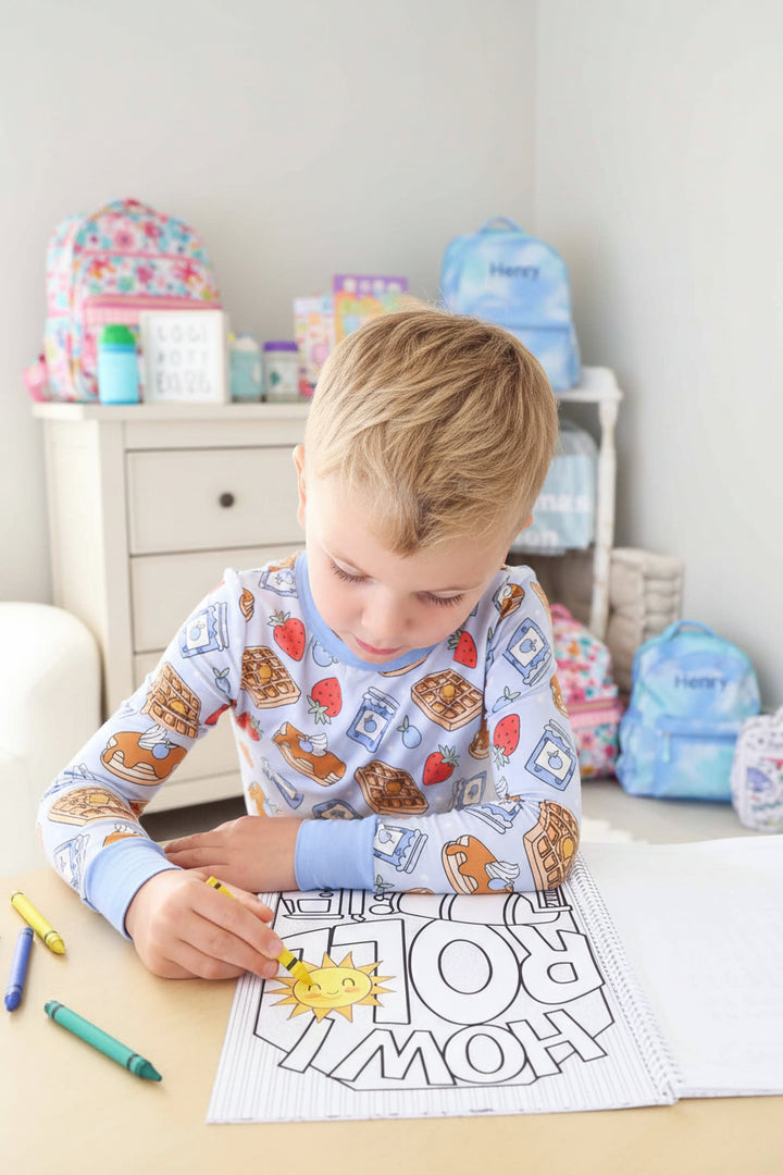 Child in a waffle-print pajama set coloring an upside-down 'Roll How' construction-themed page with a brown crayon in a Caden Lane coloring book.