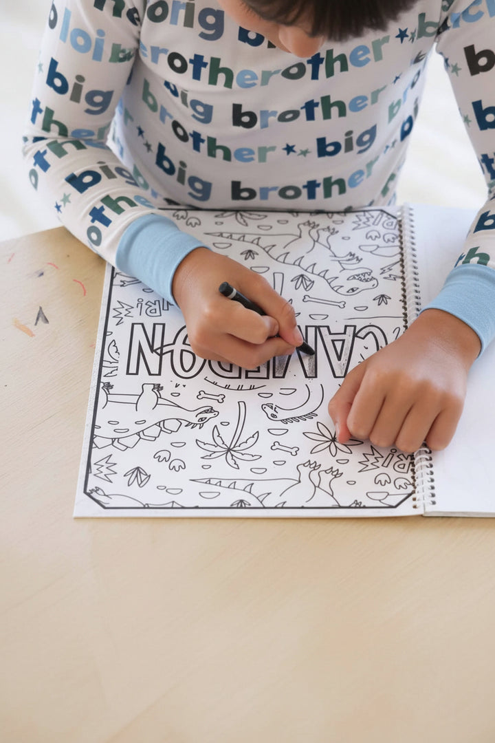 Close-up of a child in a Big Brother pajama set coloring in the letters of 'Cameron' on an upside-down dinosaur-themed coloring page with a black crayon