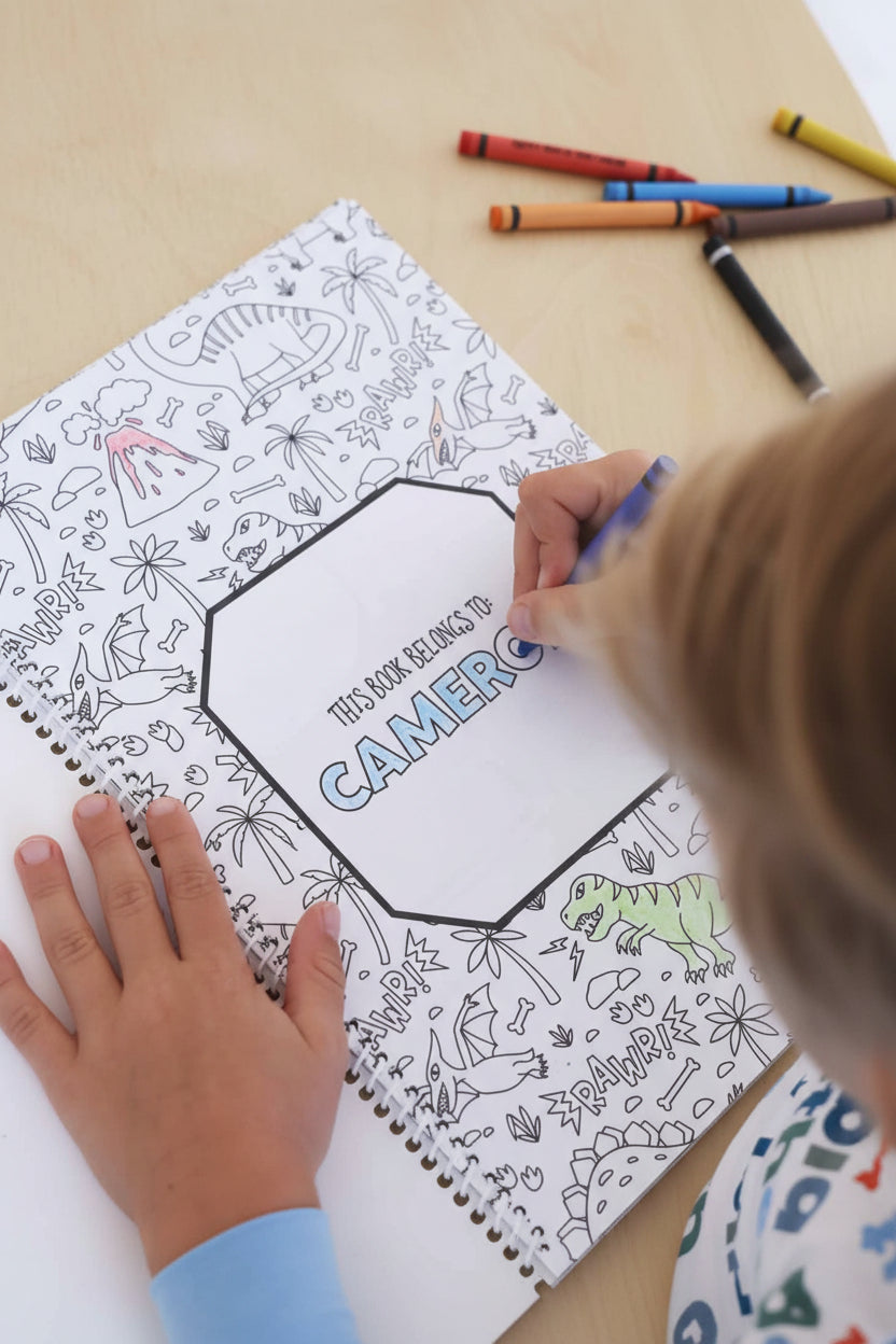 Child in a Big Brother pajama set coloring the 'This Book Belongs To: Cameron' title page with a blue crayon, surrounded by dinosaur and volcano illustrations in a Caden Lane coloring book