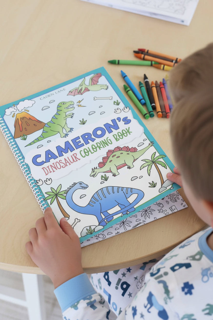 Child's hands holding open Caden Lane 'Cameron's Dinosaur Coloring Book' with a T-Rex, stegosaurus, and pterodactyl cover illustration, crayons visible nearby.